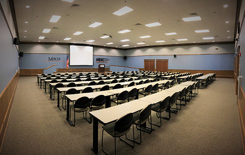 a large room set up as a classroom or auditorium with desks and chairs pointing towards a podium and projector screen